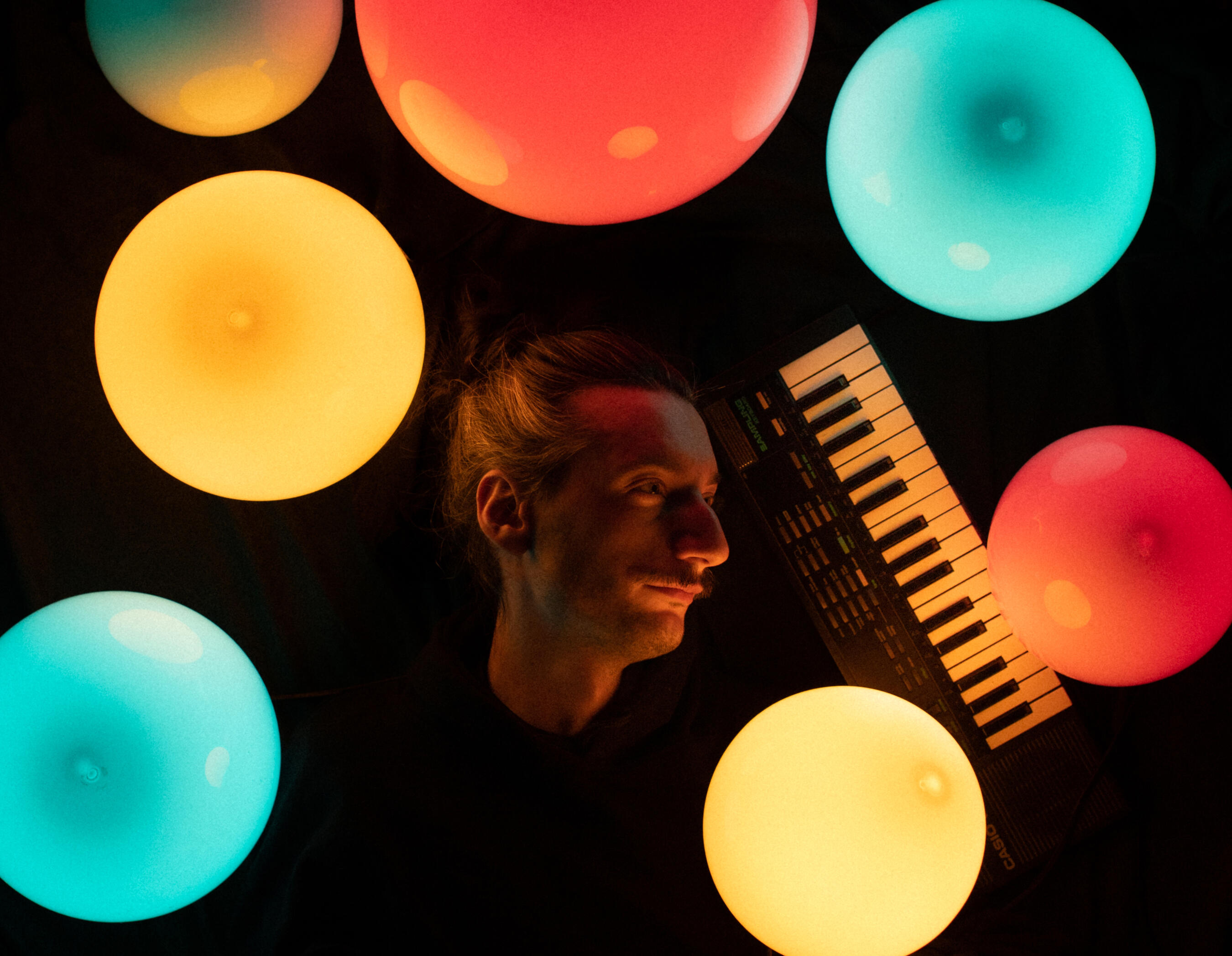 9EYE - artist pic Overhead portrait of a musician lying in darkness beside a small keyboard, surrounded by glowing colorful orb lights in red, teal, and yellow.